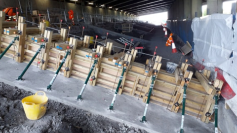 Wooden planks have been added to the bottom of a bridge. The wooden planks have been shaped into a box. The boxes have small green poles supporting their shape. A high-vis jacket and a yellow bucket have been left on the construction site. 