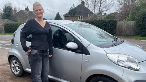 Young woman wearing a black v neck long sleeved top standing by a silver car and smiling.