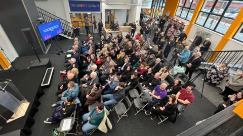 An aerial view of a large group sitting on fold up chairs facing a small stage inside the library