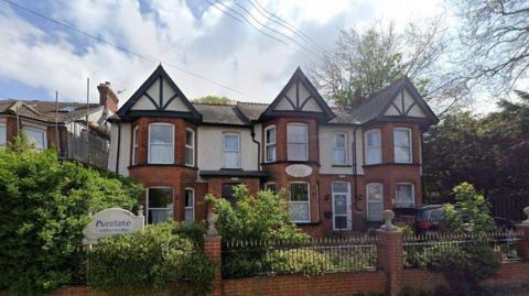 An exterior view of the suburban property, a large red brick building with the upper storey pebble dashed, white PVC window sills and shrubs and bushes in full bloom in the garden, behind a set of Victorian iron railings.