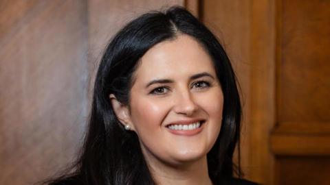 Shows a woman in long black hair smiling. A pearl earring can be seen in her right ear.
