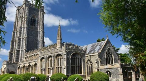 A view of St Peter and St Paul's Church church on a sunny and cloudy day. Graves can be seen in the graveyard and there are some green bushes and trees.