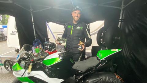 Aran Sadler stands behind a green and white motorcycle holding two trophies. He has black hair and beard and is wearing black biking gear and a baseball hat