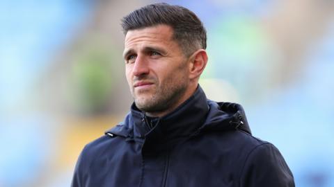 John Mousinho, wearing a black coat, standing on the sideline in the 5-1 loss at Coventry 