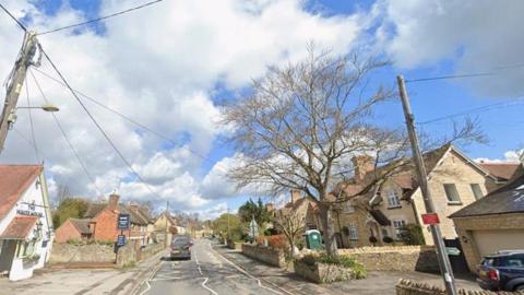 Road through a village with a white pub on the left and homes to the right.