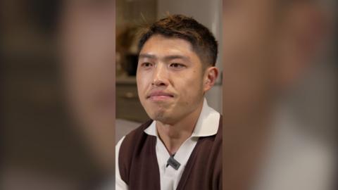 A young man looks into the distance. He is wearing a white polo shirt with a brown vest over it.