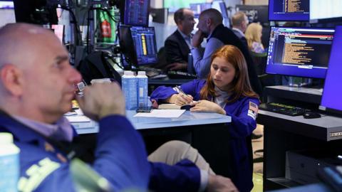 In the left corner, a bald male trader, and a woman with auburn hair, also a trader, in the middle, holding a pen and writing into a notebook, on the floor of the New York Stock Exchange. 