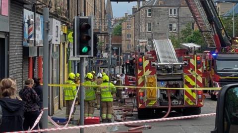 A busy street cordoned off with several fire engines and firefighters in the midst of tackling a fire