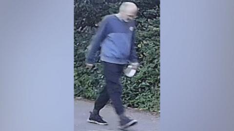 A CCTV image of an unidentified man walking in the village of Brantham. He wears a blue jumper, black trousers and black shoes.