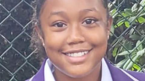 Mia Shay St Hilaire, a 12-year-old in school uniform of a purple blazer and white shirt. She is standing in front of a chain-link fence and is smiling.