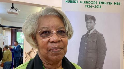 An older woman with short grey hair and darkly tinted glasses stands in front of a sign with a black and white image of a soldier. Above the full-length photograph is text reading Hubert Glendore English MBE 1926 - 2018. 