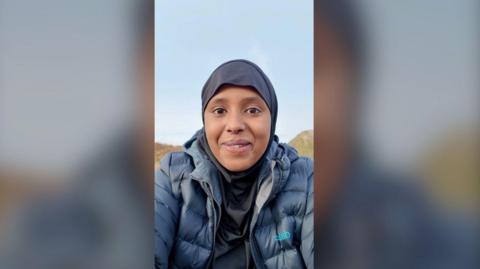 Amina Hassan smiles at the camera. She is wearing a blue rain coat, and a black hijab. There are hills behind her and a blue sky.