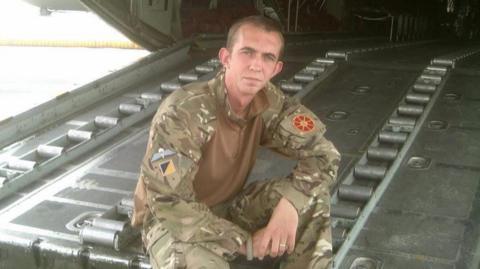 Cameron Alexander in his Army uniform sitting on a large metal door of an aircraft.