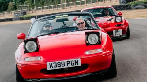Two red Mazda sports cars with pop-up headlights on a race track. Two people can be seen in the first car, which has its roof down.