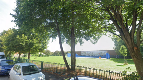 St James' Park. The picture is taken from the street, with a line of cars parked next to the pavement. There are trees lining the edge of the small park, which is fenced in, and a basketball court with blue fencing is visible on the other side of the grass. 