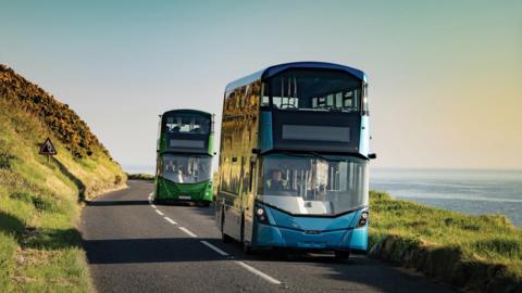 Two buses driving along a country road with the sea in the background. The first bus is blue and the second is green.