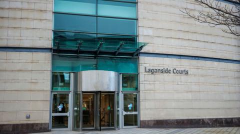 A cream brick building has a large green-transparent glass entrance with a revoling door. 