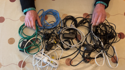 A pair of man's hands are at the top of the picture resting on a tablecloth and holding a tangled mess of cables, chargers, leads and plugs which are in a big pile in front of him. We are looking down at the scene with a bird's eye view and can only see the man's hands.