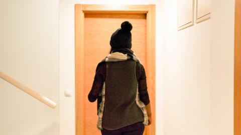 A woman stands in front of a door and wears a beanie hat.