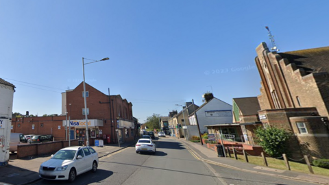 Street view of Oundle Road with a church building on the right and the plot of land on the left and cars on the street