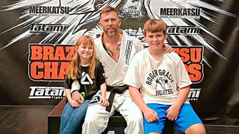 A man wearing a white gi with a black belt, smiles and has his his arms around his two children.