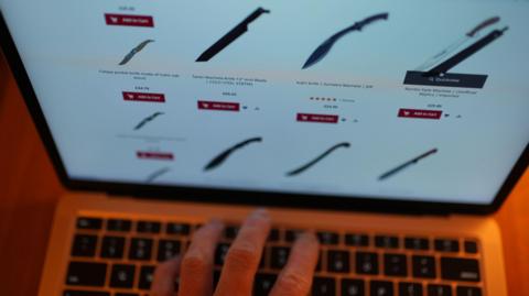 A hand on a laptop keyboard, with a screen showing a display of large knives for sale