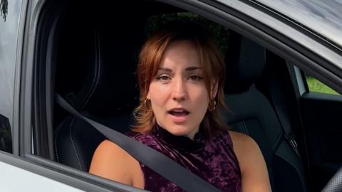 Girl with brown eyes and ginger hair wears gold hoop earrings and a crushed velvet purple vest top and sits in a grey car with her seatbelt fastened.
