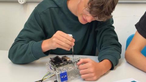 A teenage boy in a gark green jumper with short brown hair and glassed works witha screwdriver on a small platic and metal appliance.