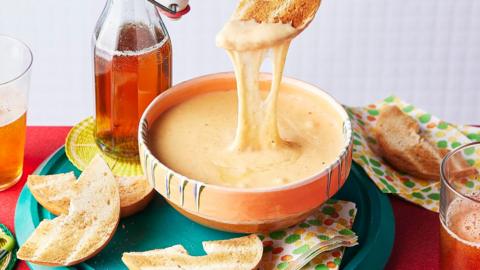 An orange bowl of a gooey cheese dup, with a chunk of bread being dipped and a glass of beer