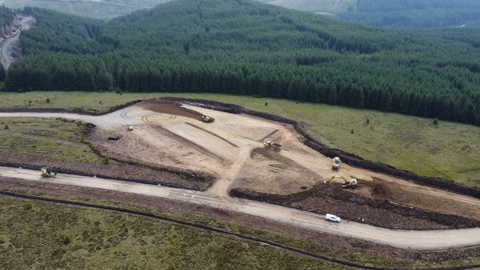 Drone shot looking down on a construction site. A number of diggers are on site on an area already dug up, surrounded by fields and trees 