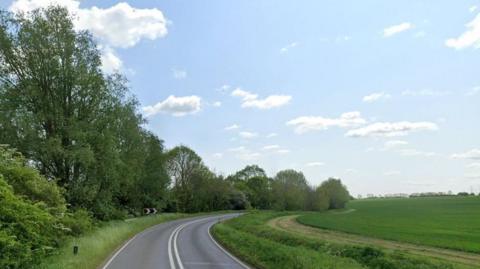 A single carriageway road with trees in leaf on the left and green fields on the right