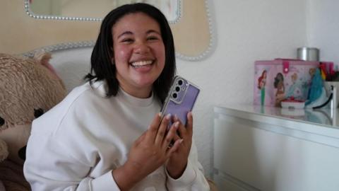 A teenage girl clutches her phone which has a purple phone case and smiles while sitting on her bed in her bedroom.