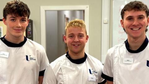 Three male students stand shoulder to shoulder. They are dressed in matching white Ulster University scrubs with a navy blue trim and white name badges. From left to right, they have dark wavy hair, light blonde hair and light brown wavy hair. They are in a classroom environment with a whiteboard and door behind them. 
