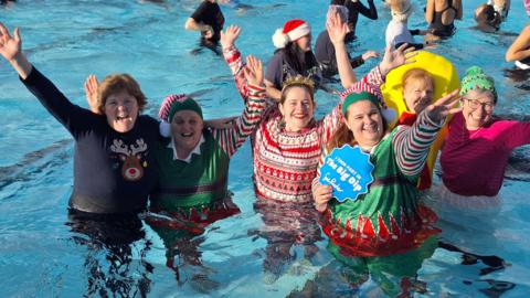 Six women are pictured standing in an outdoor pool wearing Christmas clothing. One is dressed as an elf, another in a Christmas jumper with Christmas tree earrings. They are standing and smiling with their arms in the air. 
