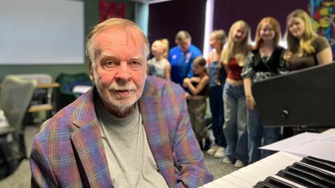 Rick Wakeman stand at a keyboard with a line of girls in the background.
