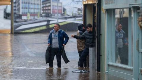 Three men stood outside a shop on Mill Street in Pontypridd, wading through flood water. One, wearing a blue coat, is stood in the street. Water can be seend to be entering one property on the right hand side.