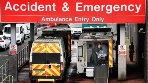 Two ambulances parked outside an A&E department. One has its rear doors closed while the other has its doors open. A bald paramedic, wearing green overalls, can be seen adjusting a bed in the rear of the vehicle.