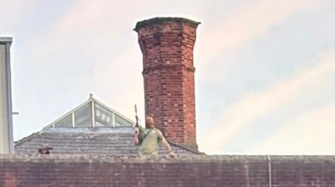 The blurred shape of a man in a green top holding what appears to be a rifle on the roof of a brick building with a large brick chimney behind him