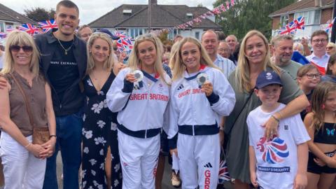 Kate and Isabelle wearing Team GB tracksuits wearing their silver medals, surronded by their family, friends and neighbours, outside on their cul-de-sac