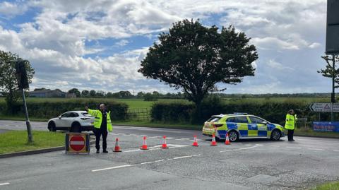 Two police officers in hi-vis uniforms at the scene of the crash. There is a police car blocking the road and cones in place along the road. There are trees and green field in the background.