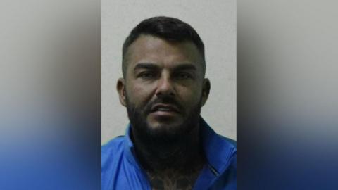 Mugshot of Luke Gallagher. He is heavily fake tanned, has straight white teeth and a short dark beard and hair. He is wearing a blue jacket which is open at the front revealing an enormous tattoo of shapes and patterns on his chest.