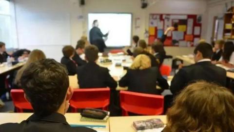 Secondary school children in a classroom from behind, they are wearing black blazers and sat on red seats, a teacher points at a projector screen.