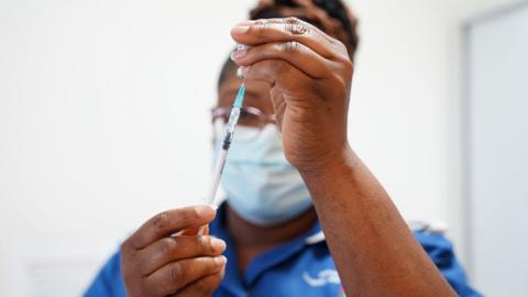 A close up shot shows a nurse holding a syringe inside a vile while preparing a Covid booster jab, she's wearing a medical facemask and blue nurse outfit against a white background. Picture taken at University Hospital Coventry in 2022.