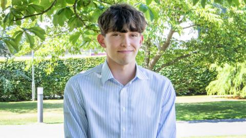 Felix Merritt wearing a blue and white pinstripe collared shirt and smiling at the camera. He has thick brown hair which covers his forehead, and is standing beside a concrete path below trees and low-hanging branches. It is a bright sunny day and the image looks warm.