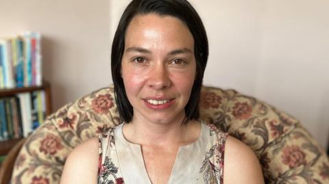 Stef Benstead with shoulder length brown hair and brown eyes is sitting on a chair with a floral pattern wearing a beige top and smiling. 