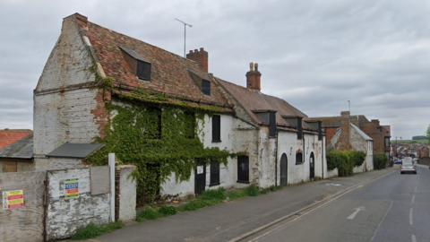 A derelict three-storey building next to a road in Newmarket. It is overgrown with vegetation and weeds and looks in a poor condition.