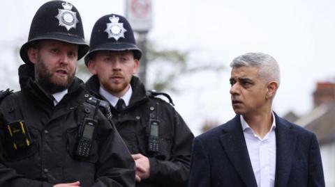 Sadiq Khan is seen on the right of the image walking next to two make police officers. His head is turned towards as he appears to be listening to him.