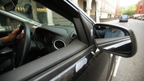 A driver viewed through the wing mirror of his black car is shown not to be wearing a seat belt