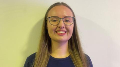 A woman with glasses and long brown hair is in front of a yellow and white background. She is wearing a blue T-shirt. 