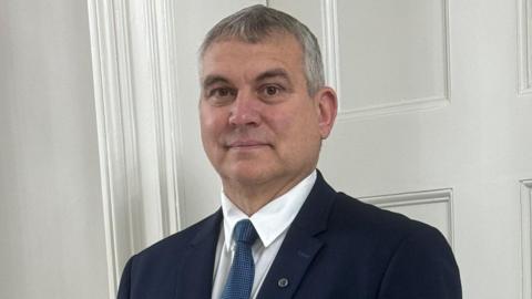 A headshot of a man with grey hair wearing a navy suit, white shirt and blue tie smiling into the camera. He is standing in front of a white wall. He has a small badge on his lapel. 